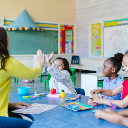 photo of young kids with their teacher. One student is high-fiveing the teacher, the other student's watch and do crafts.