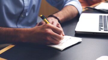 person writing at a desk next to laptop