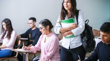 Student standing and holding books in the middle of a classroom of students sitting at desks