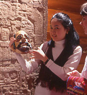 A young girl holds a mask. In the background is a carved stele. 
