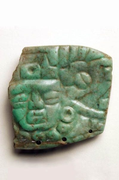 Polished piece of greenstone with a carved face above three small holes.
