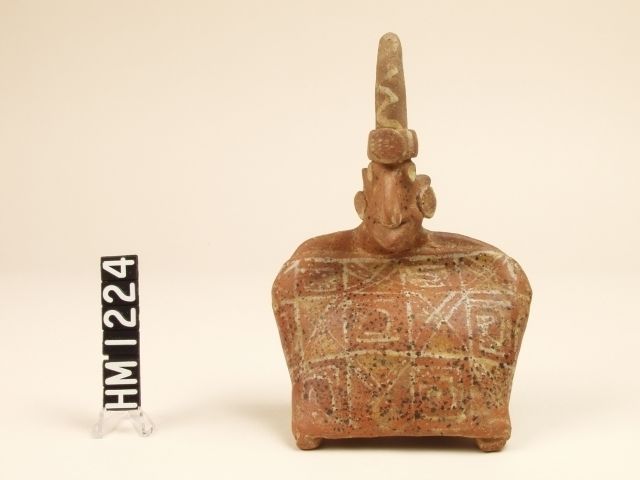 Small ceramic figure. Body is a squat rectangle with a head at center top. Large, pointed headdress.