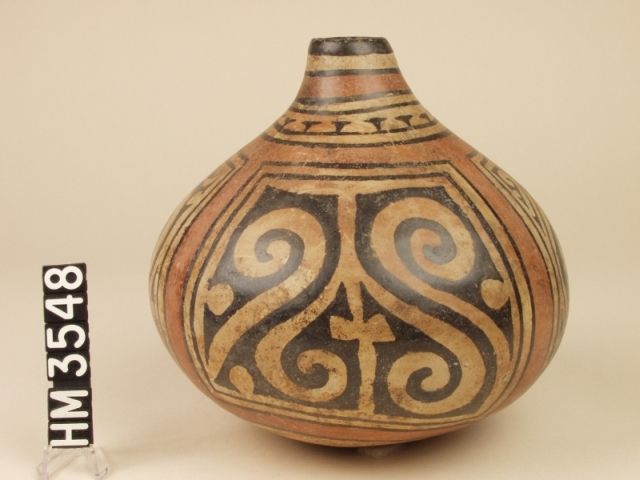 Polychrome ceramic bottle. The side panels depict claws. The front and back panels use Y-elements to suggest an abstract frontal face or crested head. The face is formed by two pointy-nosed crested animals in profile, facing each other. The crest is formed by the tail-ends of the profiled creatures.