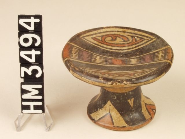 Miniature polychrome ceramic pedestal plate. The design on the top of the plate features a central bar as a serpent with boa ovals and rectangles represented by alternating red and cream bands. Adjacent hemispheres each contain a "Y" element with a claw or a curly crested "Y" element head and tail. In either case, an animal kenning is encased within boa ovals.