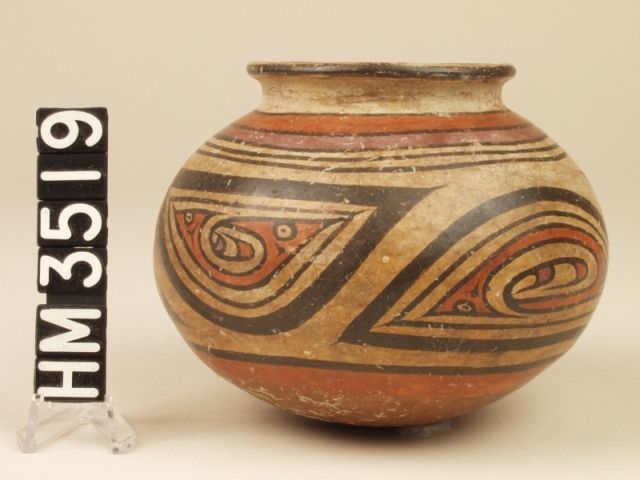 Miniature polychrome ceramic jar. A red "Y" element design inside the black border of an oval panel depicts a profiled creature with a pointed nose, a curly head crest and a horizontal eye slit. A tail end in the form of a second head is also indicated by a slit eye. A single basic "Y" element forms the overall body, and the tail (head) end and crested head end are crested by additional "Y" elements.