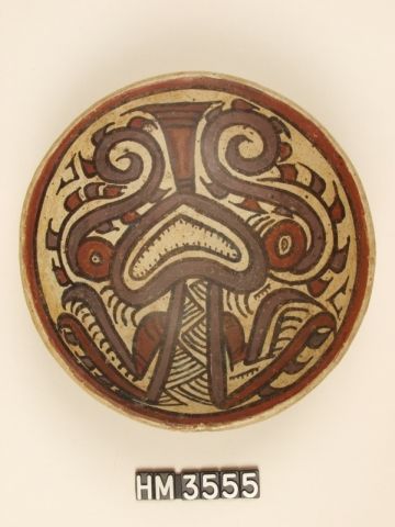 Polychrome ceramic rounded bowl seen from above. Color contrast defines the depicted creature as a composite iquana-boa. Boa features, colored purple, include an open toothed mouth, body line and eye stripes. Boa kennings, sets of parallel lines, are also depicted in the main body. Iguana characteristics are colored red. They include the eyes, portions of limbs and a thick, protruding tongue. The positioning of the eye stripes as a type of curly head crest also suggests the theme of the iguana.