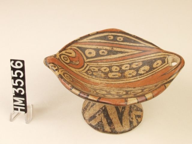 Polychrome ceramic pedestal plate. Spots and shape suggest either an eagle ray or a round stingray. The spots are broken on one half of the design by a depiction of a tail with a protruding spine. Boa-like eye stripes and banded design on the rim. One end has a hole through the clay; the other end is the eyes and nose of the animal.