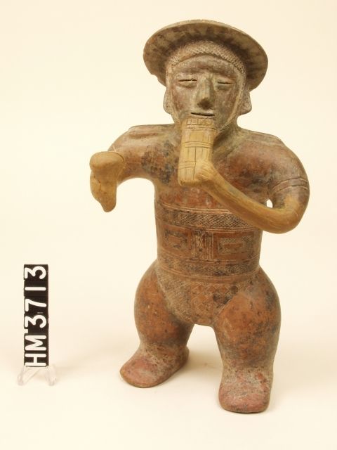 Ceramic standing figure with hat and ear spools playing reed flute with left hand and holding rattle in right hand.