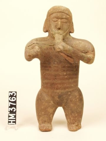 Ceramic figure of a standing man playing reed flute with left hand and holding rattle in right hand.