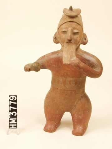 Ceramic standing figure with headdress playing reed flute with left hand and holding rattle in right hand.