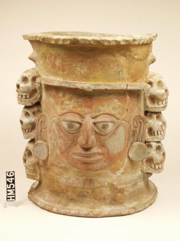 Ceramic vase with vertical sides. A face is painted on the front. Six skulls flank the face, three on each side, attached to the sides of the vase.