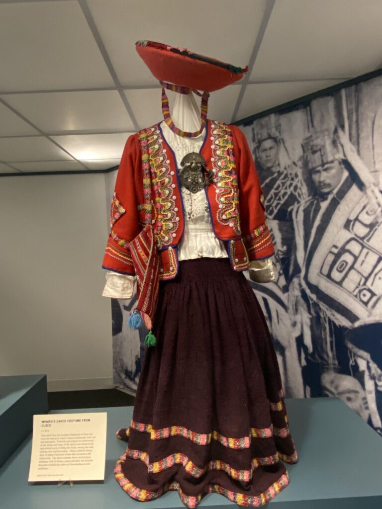 Image of an outfit on an exhibit stand. It includes a funnel-shaped hat, intricately-embroidered red jacket, woven bag, and long skirt with three decorative strips at bottom.