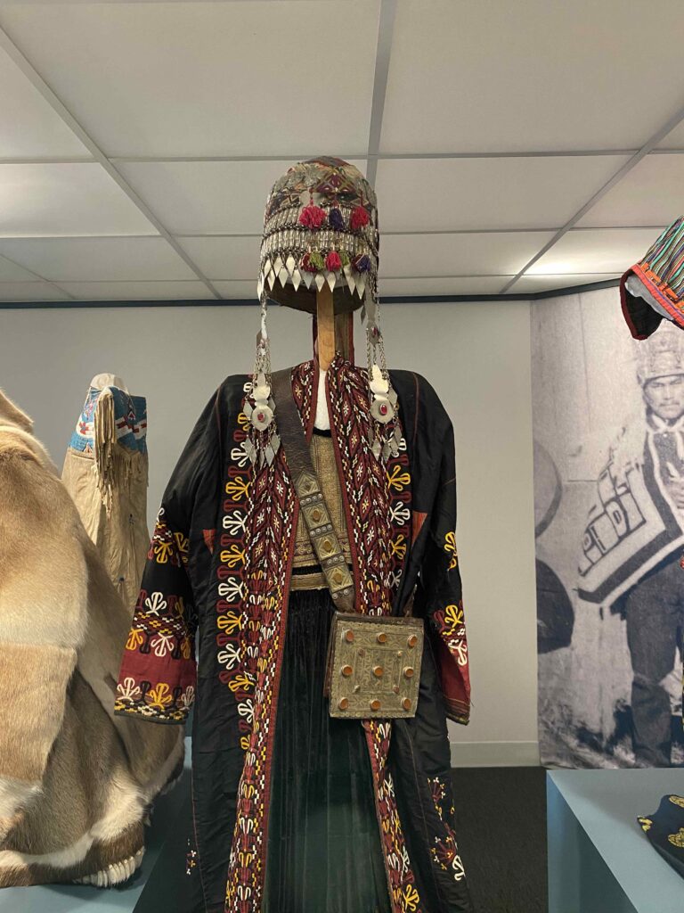 Image of an outfit on an exhibit stand. It includes an intricate hat with silver elements along edges and at sides, a long intricately-embroidered coat over a long skirt and a bag with silver embellishments.