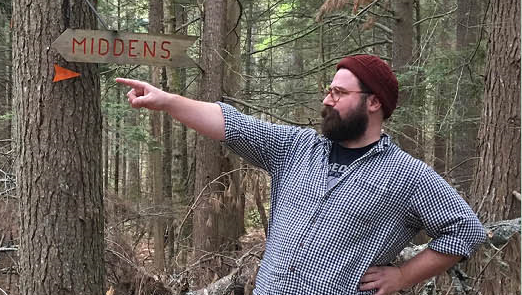 Image of a man standing in a forest. A sign behind him reads "midden" and he points in the same direction as the sign.