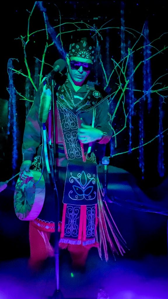A man stands in colorful light wearing an intricate costume featuring double curves and holding a drum.