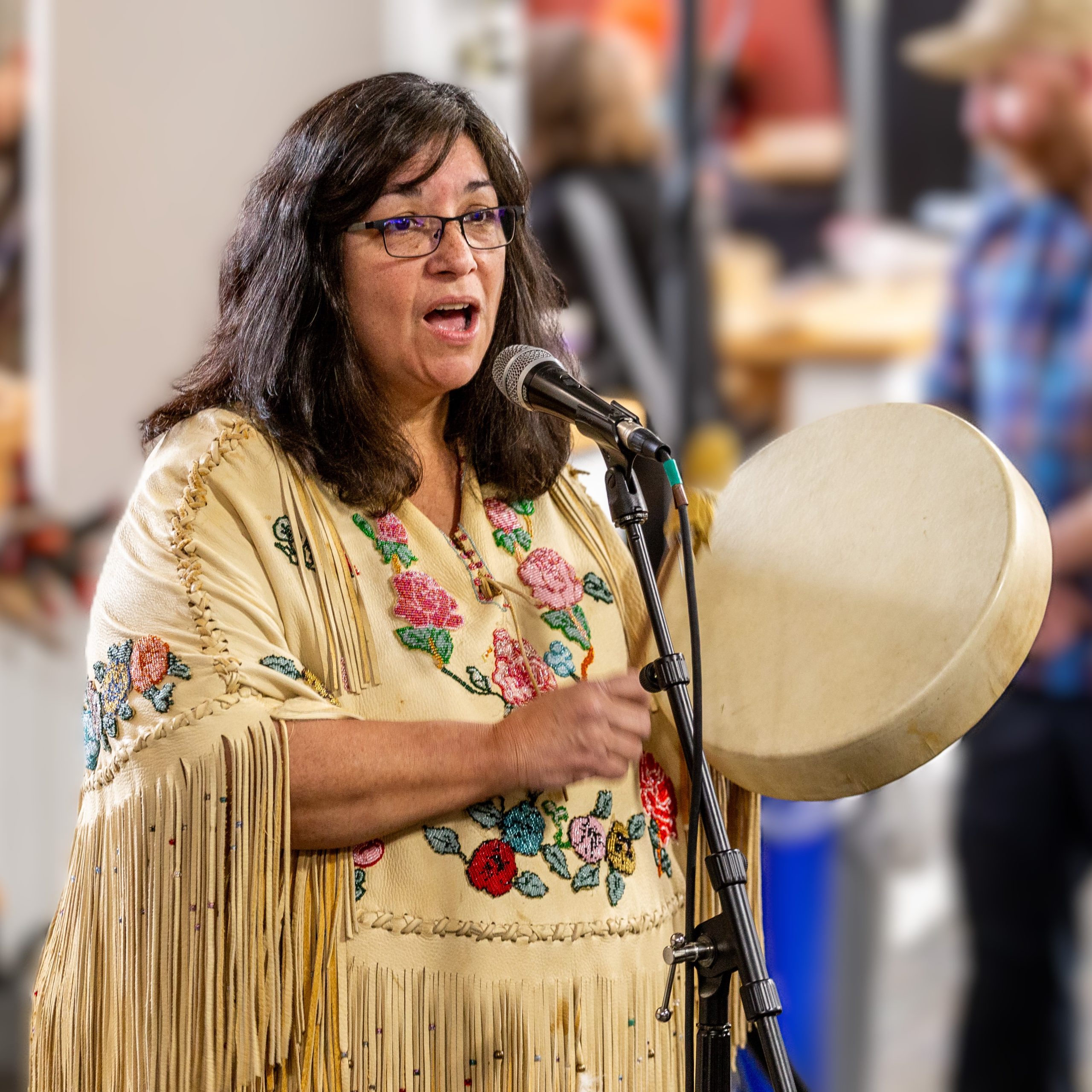 A woman in a fringed and beaded leather shirt sings at a microphone and beats a drum she holds in her left hand.