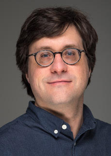 Image of Caucasian man with dark hair and small, round glasses smiling at camera.