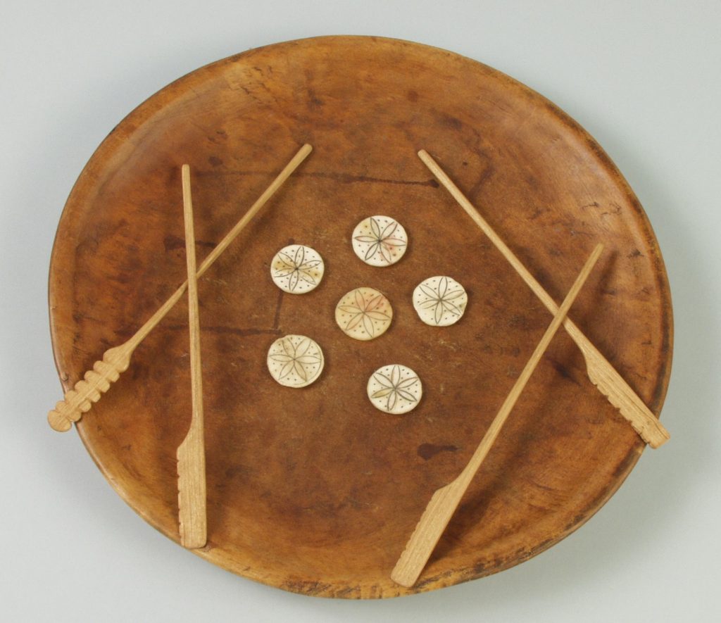 Image of a wooden bowl with five round game pieces and four long wooden pieces.