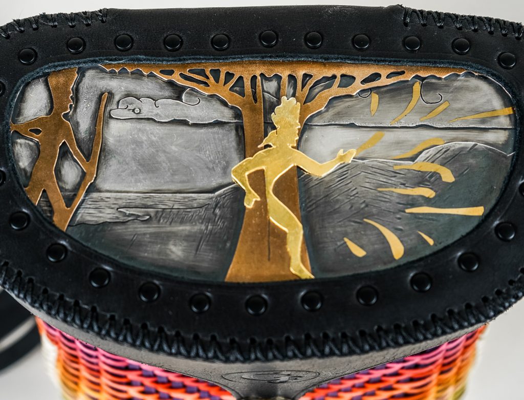 Detail of the top of a basket featuring an image made in metals. A man with a bow on the left, a man coming from a tree center.