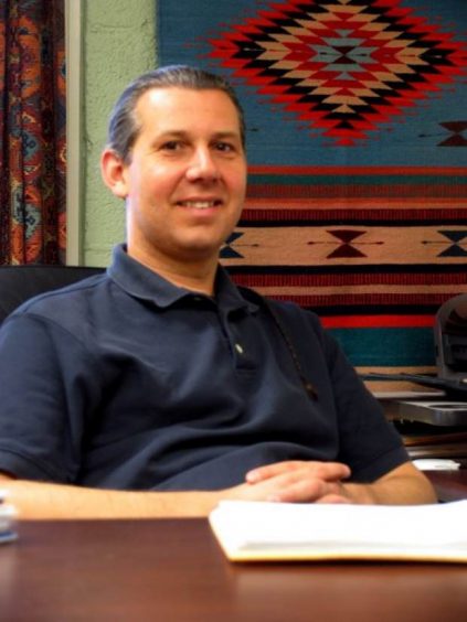 Image of Penobscot man with hair pulled back sitting at a table with a woven textile in the background