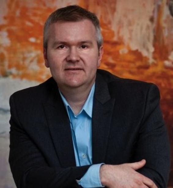 Image of Caucasian man in collared shirt and jacket with bright art in background.