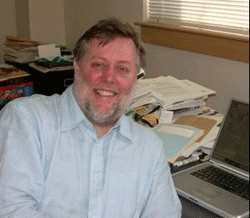 Image of Caucasian man with dark hair and gray beard sitting with a desk in background.