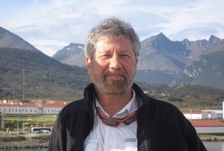 Image of Caucasian man with gray hair and beard standing with mountains in the background.