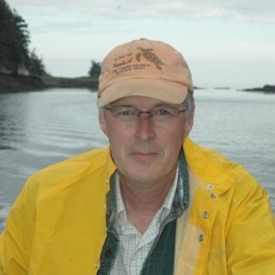 Image of Caucasian man wearing baseball cap and yellow raincoat with lake in background.