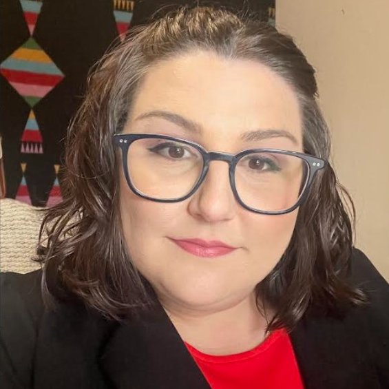 Image of Caucasian woman with dark hair and glasses wearing a red shirt and black jacket with a woven tapestry in background.