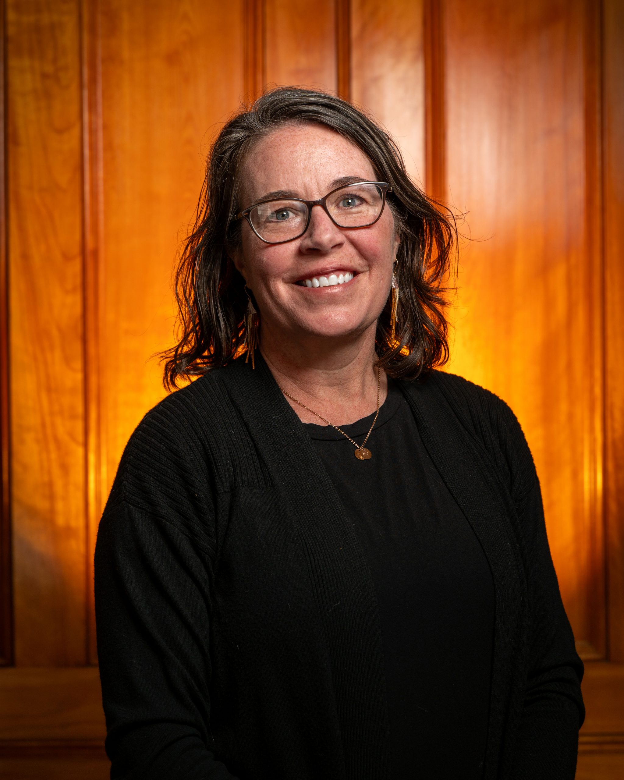 Image of Caucasian woman with glasses wearing a dark top sitting before wood paneled wall.