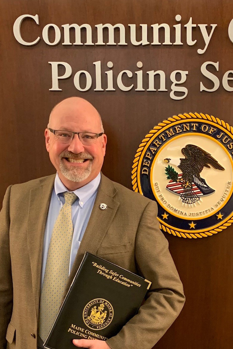 Image of bald Caucasian man standing before a wall reading "Community Oriented Policing Services"