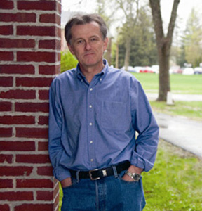 Image of Caucasian man wearing jeans and a light denim shirt and leaning against a brick wall outside.