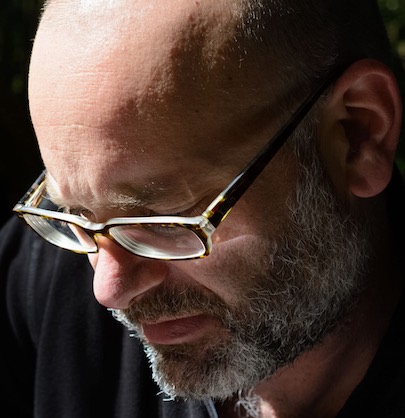 Image of Caucasian male wearing glasses looking down toward left and lit dramatically from above.