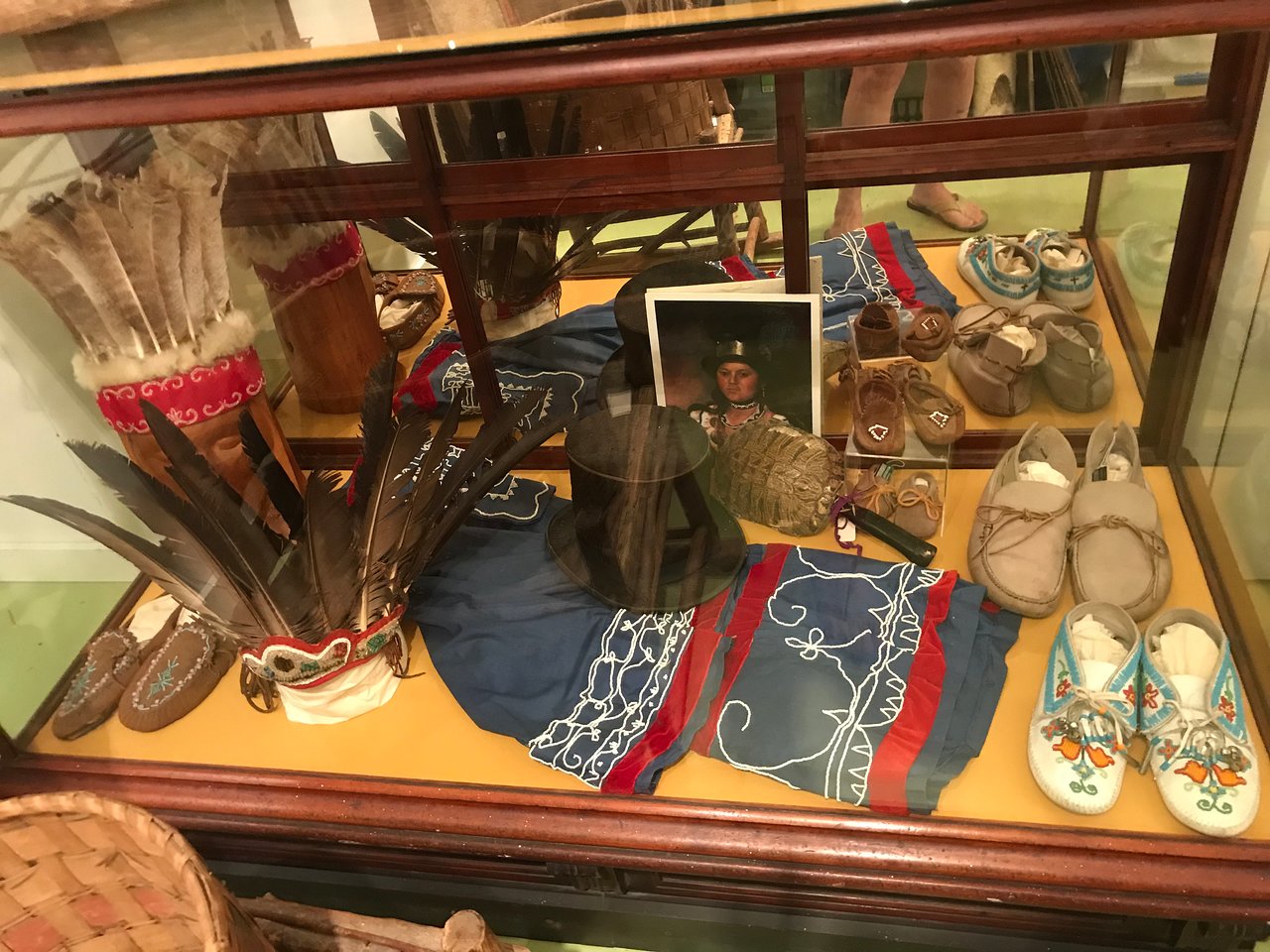 Image of a museum case with moccasins, beaded clothing, feathered headdresses, and a picture of a woman in a tophat with silver hat band.