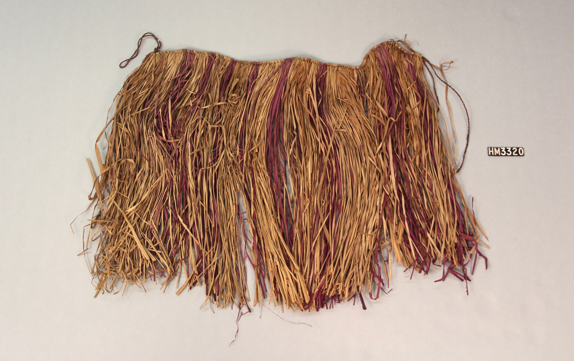 Skirt made with strands of grass attached to a string. at regular intervals along the string are bundles of grass dyed reddish purple.