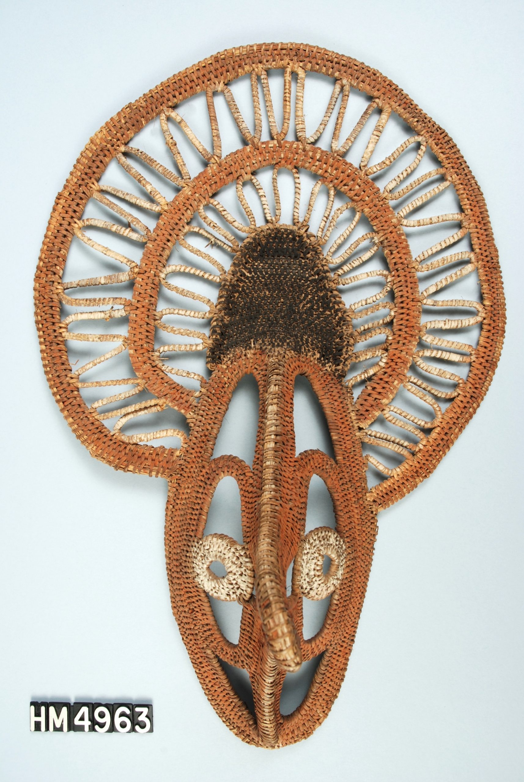 Image of mask with stylized face below large open crest, all made of woven fiber painted with orange, white, and black.