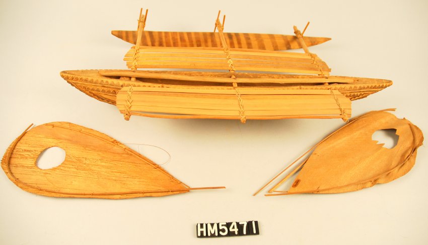 Image of a boat model with a long, canoe-shaped hull decorated with carved details. A single outrigger sits to the back. A platform sits beyond the edge of the hull on each side. Two raindrop-shaped sails are laid in the foreground.
