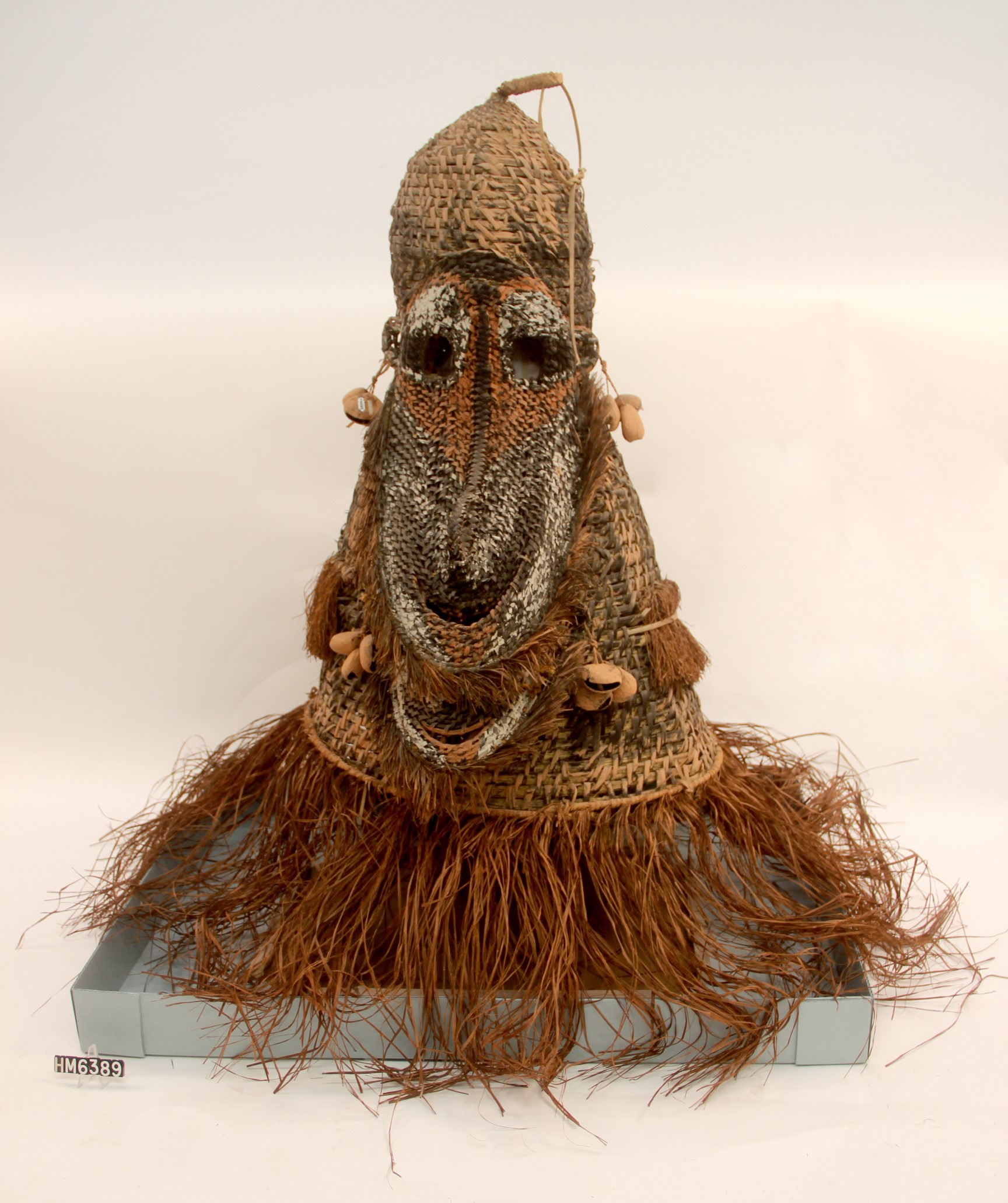 Image of a large mask made to fit entirely over the head and shoulders of the wearer and featuring a larger-than-life face and a fringe of fiber.