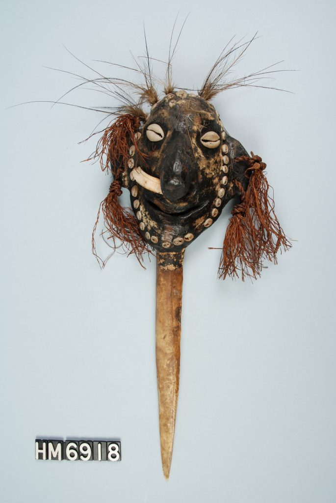 Image of an object with a long, pointed dagger made of bone below a face made of a black material with shell, feather, and fiber decorations.