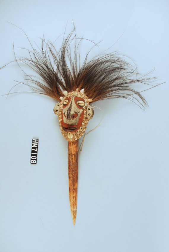 Image of an object with a long, pointed dagger made of bone below a face painted red and white with shell and feather decorations.