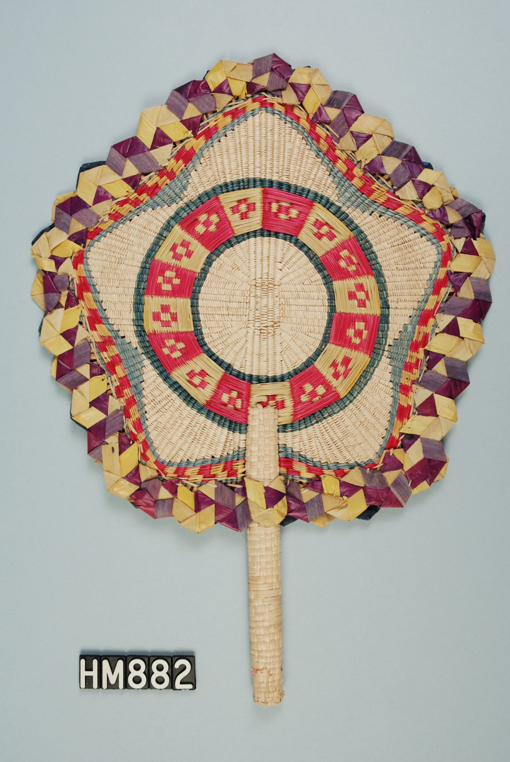 Image of a hand-held fan with circular head. Handle is woven. Body of head is woven in bands ending in one shaped like a star with five points and surrounded by a band of wider woven materials inn multiple colors.