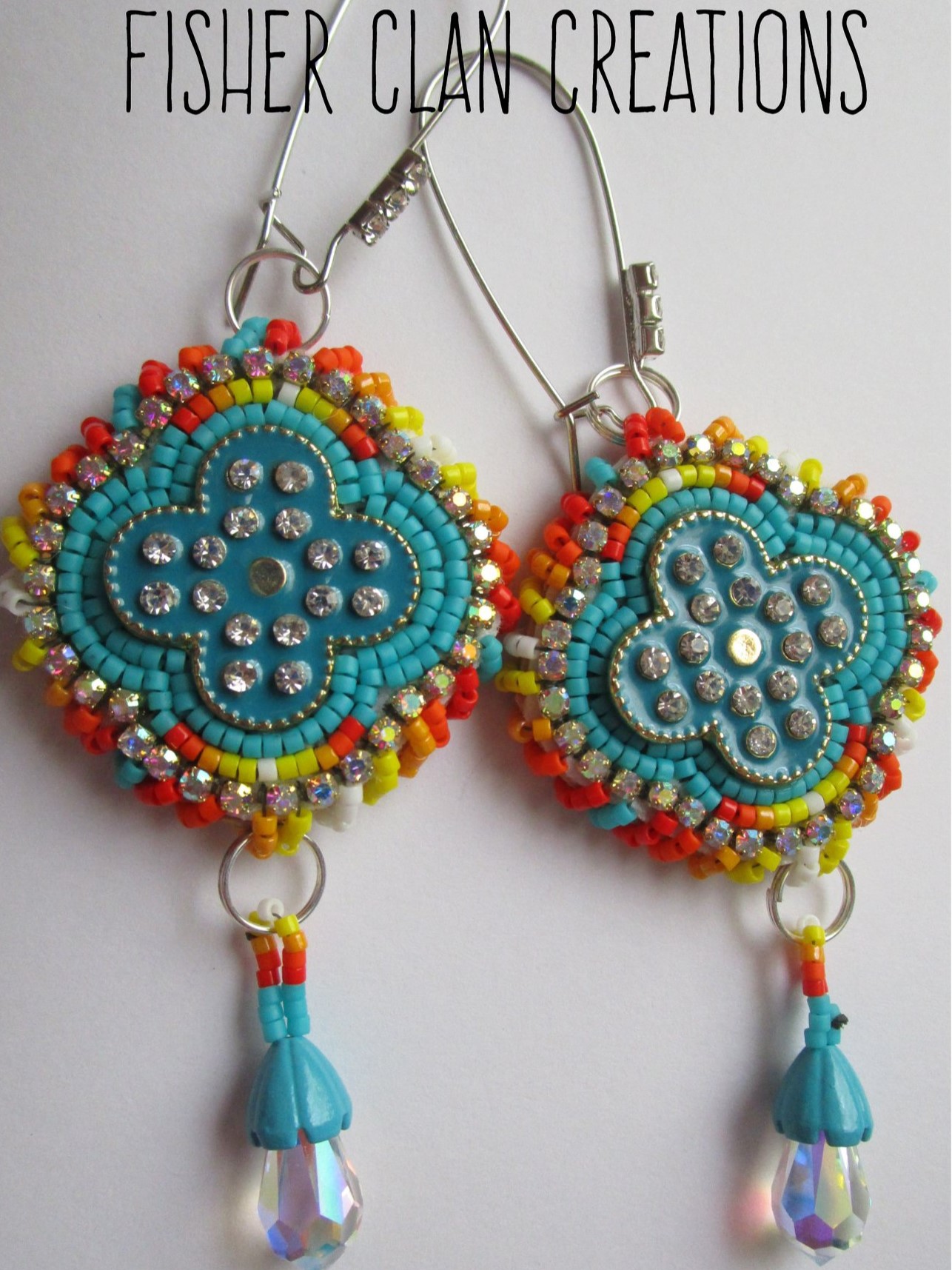  image of earrings made of dyed and beaded leather in teal with orange and yellow accents and clear gemstones. Text reads "Fisher Clan Creations"