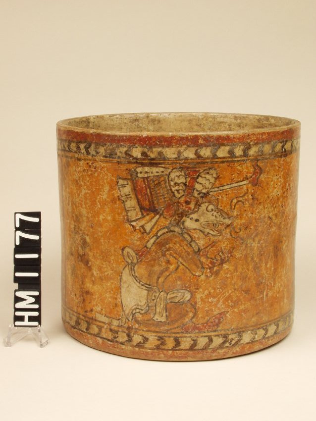 Wide vase with vertical sides. Painted with figure of anthropomorphic Fox God seated, tailor-style facing to the right and gesturing with hands in a conversational style. Elaborate headdress with grid design and scalloped lines. Chevron banding bottom and top.