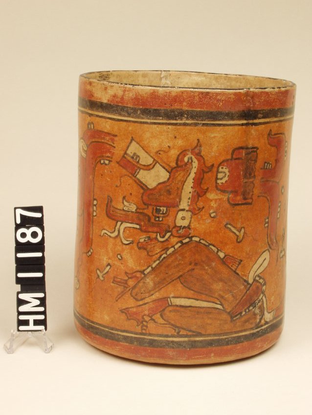 Vase with vertical sides. Red, black, and orange over cream slip. There is a traditional red rim below which is a thick black line and a fine black line. A seated, masked gesturing figure is depicted.