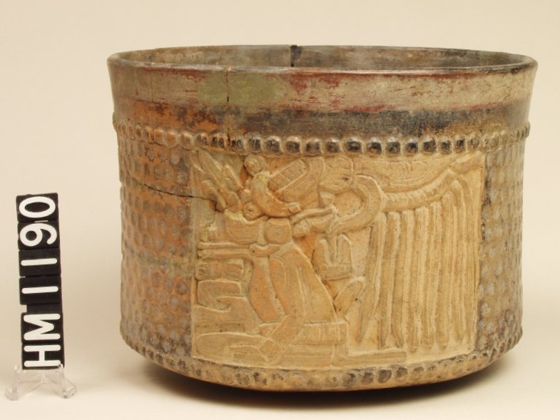 Wide vase with vertical sides. Incised with a panel of a seated god surrounded by a dimpled area. A band of raised dots encircles the top and the base of the vase. The rim is painted with a band of red above a band of black.