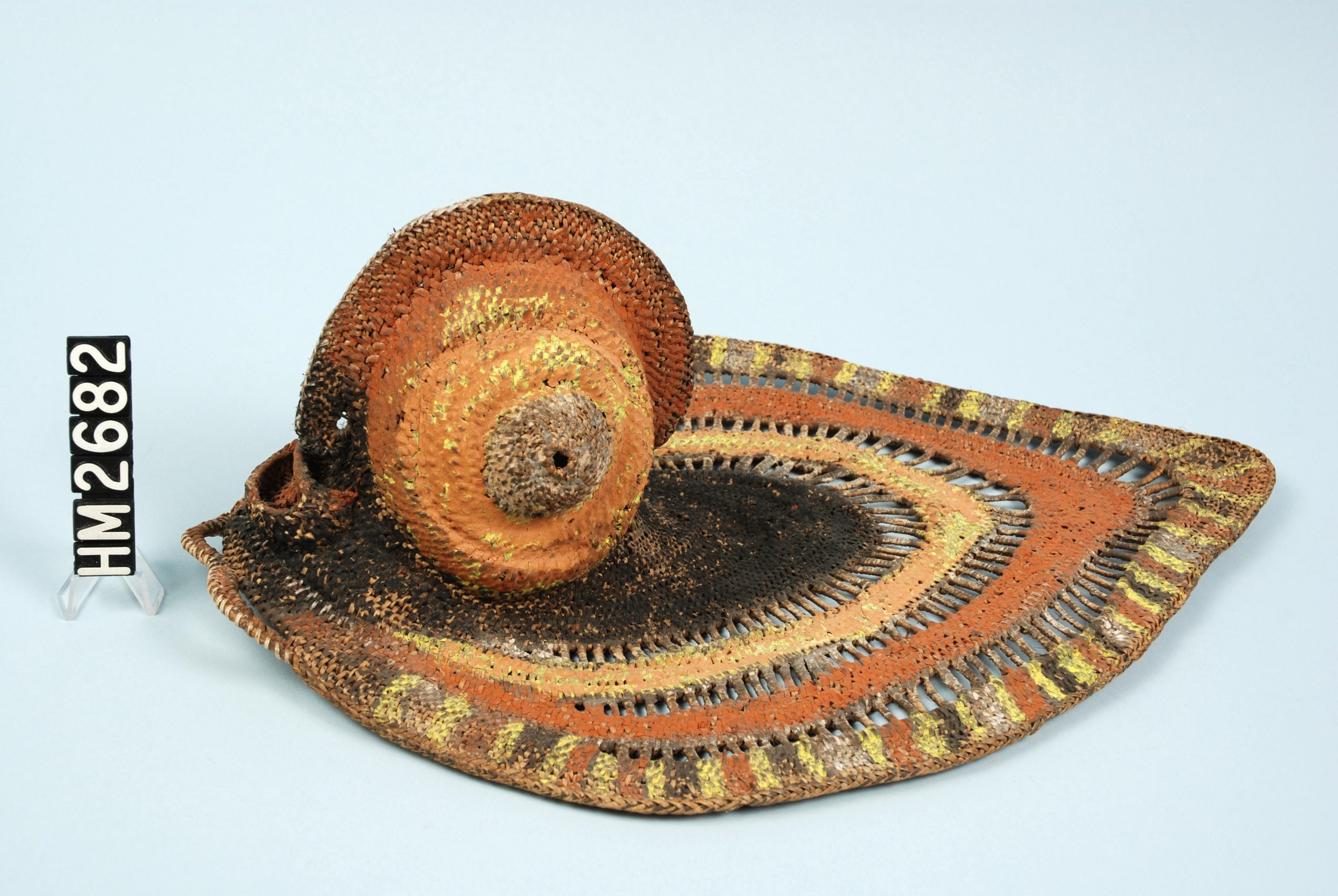 Object made of woven fiber and painted with orange, yellow, and black. In form it has a large, flat collar portion with a raised bulbous portion.