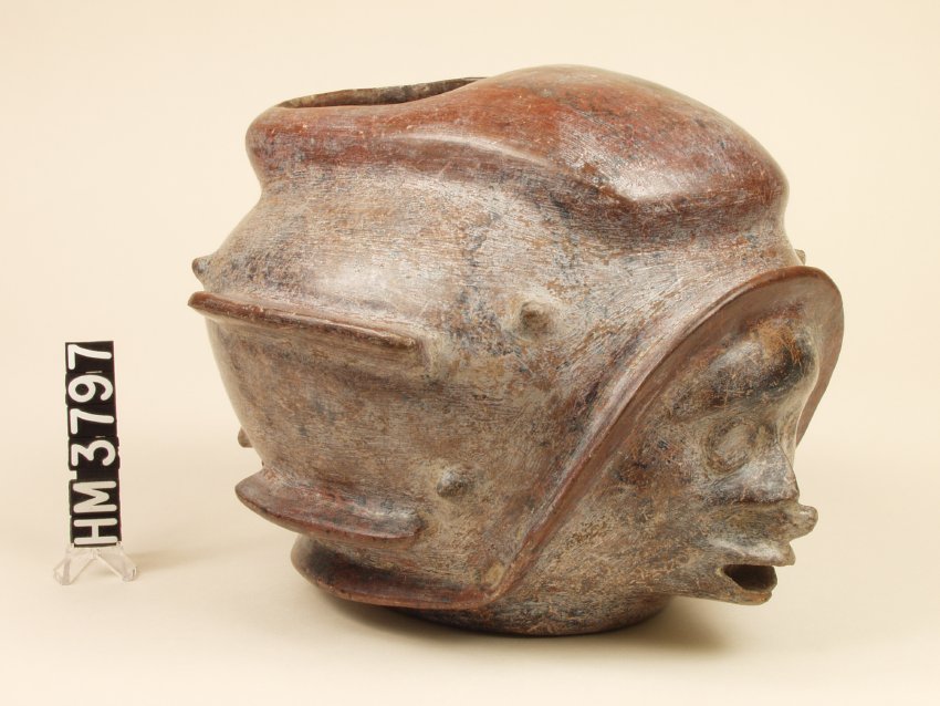 Ceramic vessel with elongated, beak-line mouth, and close-set eyes, perhaps a diminutive frog(?) on the upper lip; wearing a helmet-like headdress with fins, knob projections, and symbolic motifs.