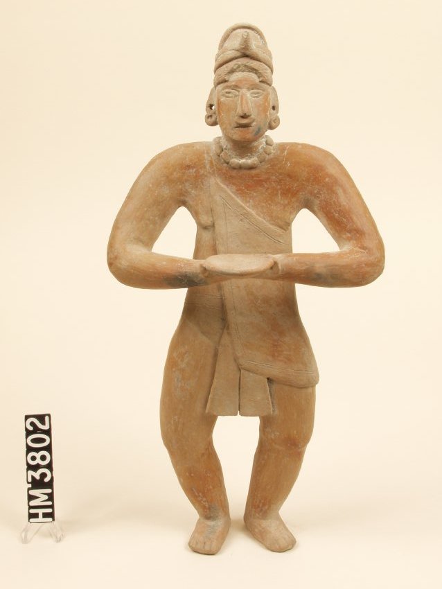 Standing ceramic figure holding offering plate and wearing a toga-looking outfit, with the cloth draped over one shoulder, a large beaded necklace, and earring on each ear.