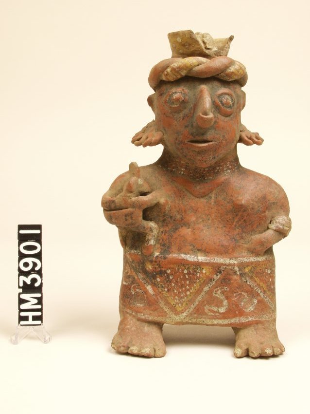 Standing ceramic figure with short arms, left flexed and hand on abdomen, right flexed and holding a child. Defined facial features, white painted eyes. Twisted horizontal headband, bowl on top of head. Wearing a skirt with white painted rim on top and bottom. Design is alternating triangles with designs in yellow and white, bead-like necklace.