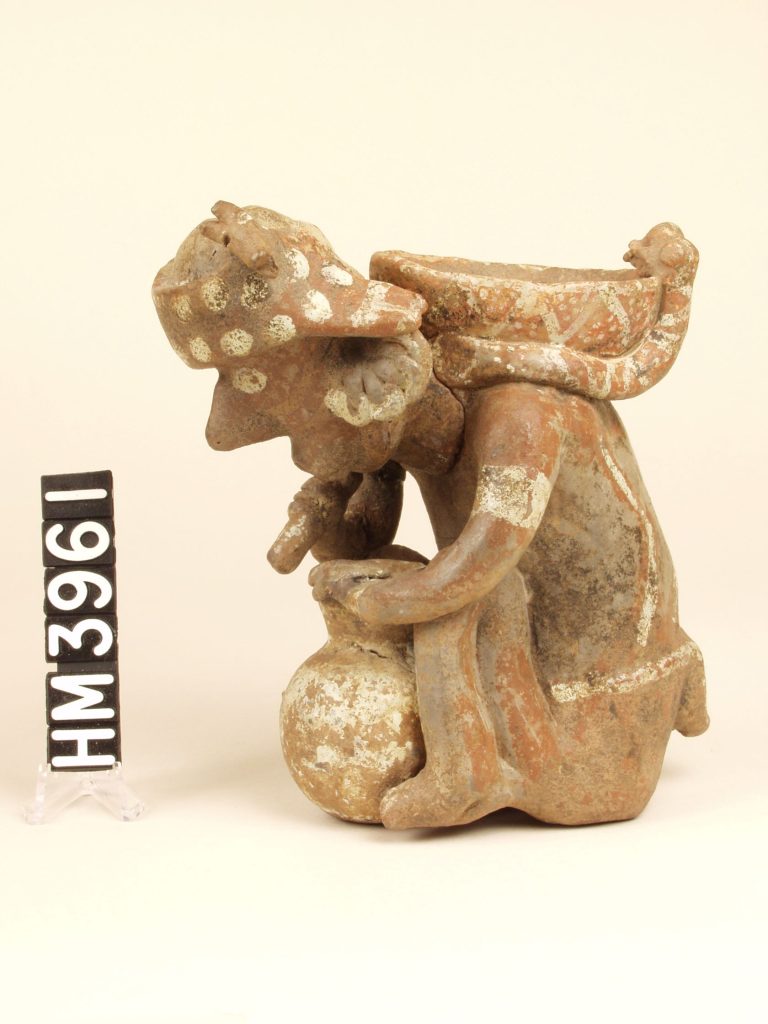 Small ceramic figure seen from the side sitting with jar between knees. Shallow bowl on back of figure, decorated with chevrons. Two snakes encircle the bowl.
